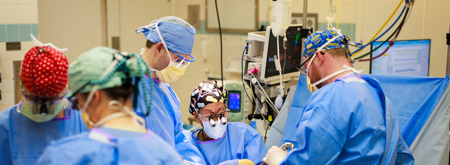 Medical team in surgical gowns and masks performing an operation in an operating room with monitors, surgical lights, and instruments in use.